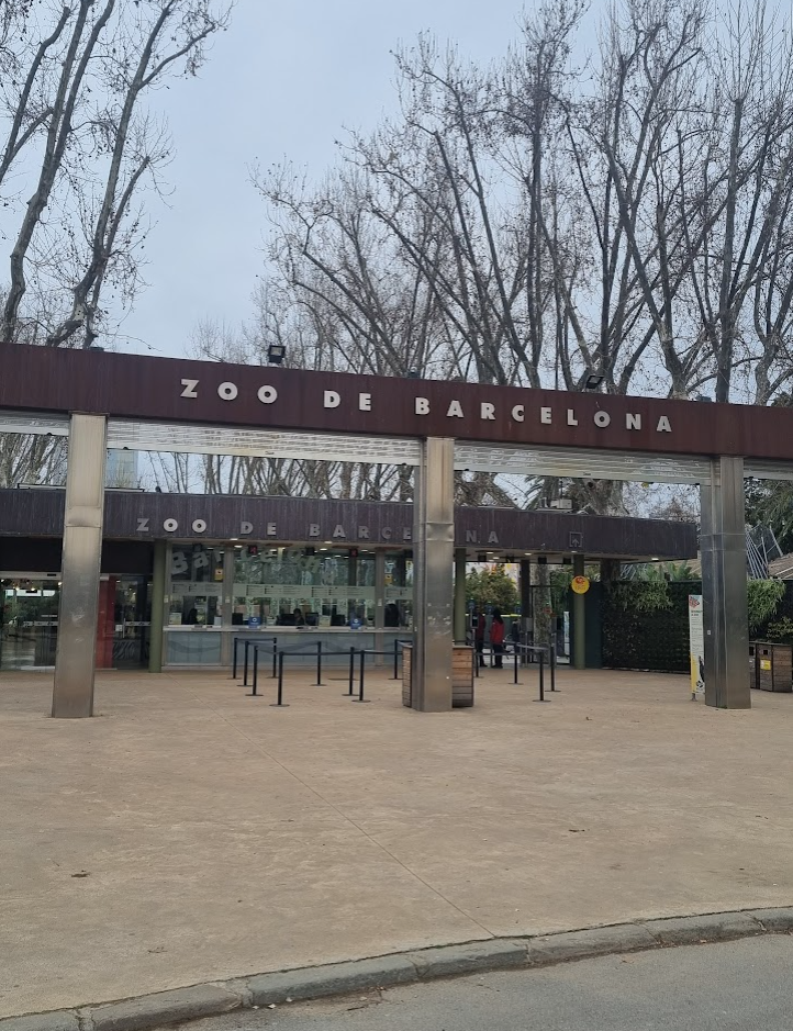 Entrance to the Barcelona Zoo at Ciutadella Park, with a sign displaying the zoo's name and logo, surrounded by lush green trees and bushes.